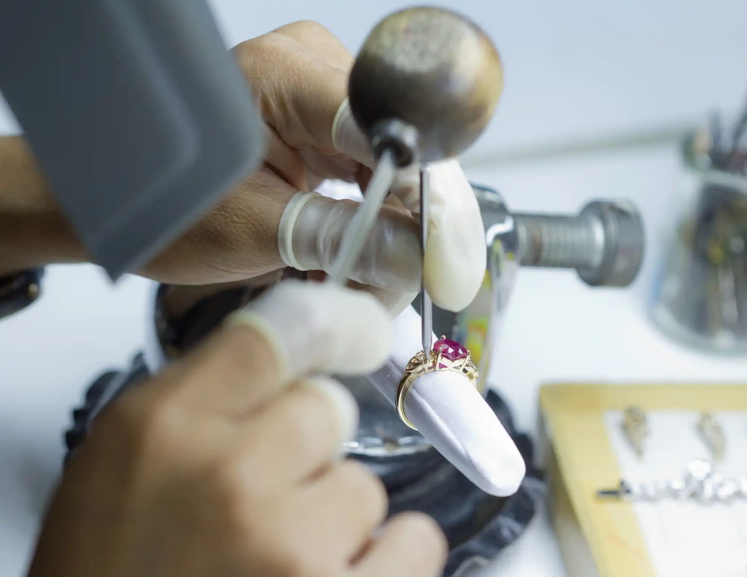 A gold ring with a prominent red gemstone, likely a ruby, surrounded by smaller, clear stones, being meticulously set or polished by gloved hands using specialized tools.